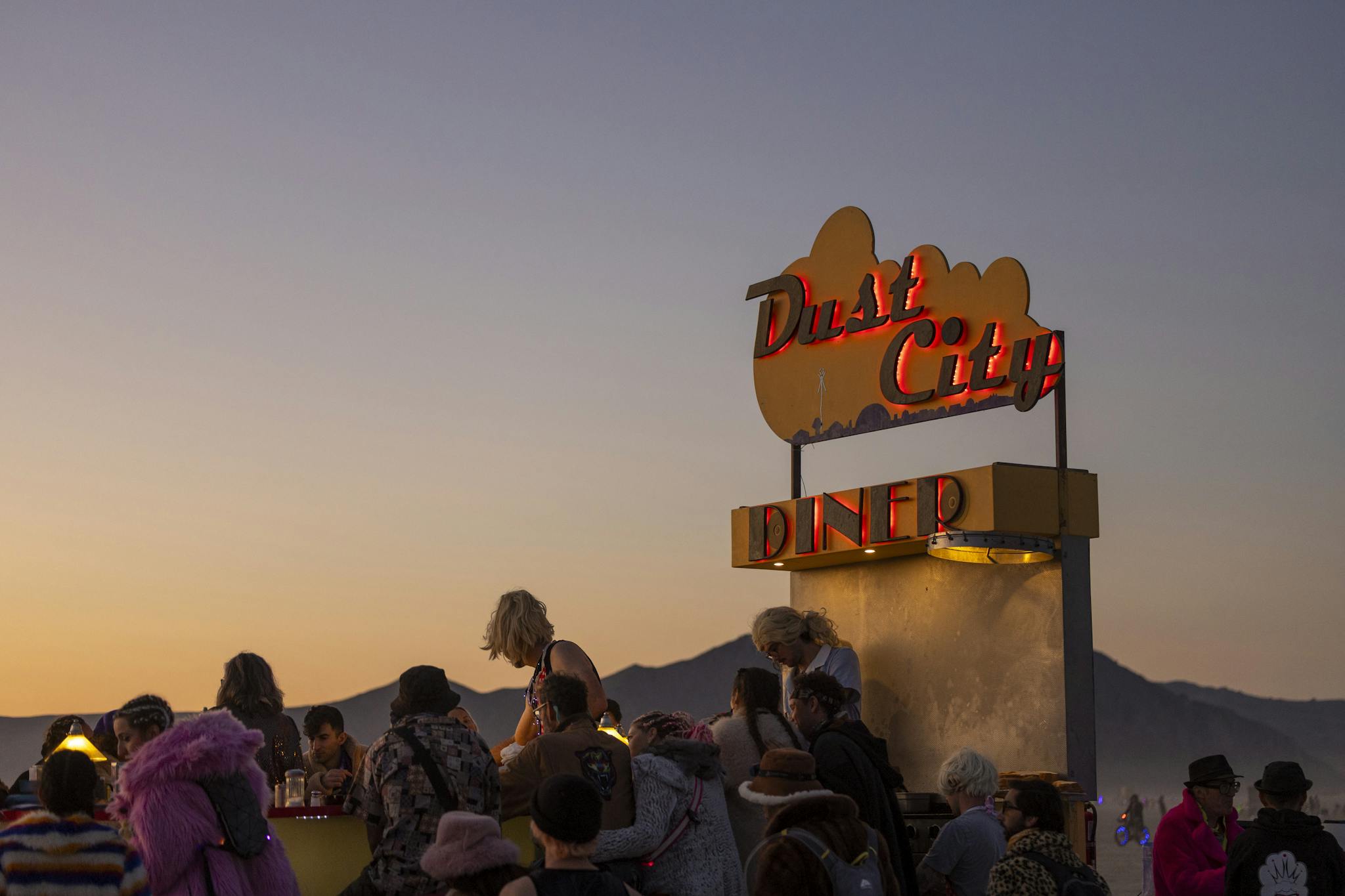 A lively group gathers at Dust City Diner during sunset, surrounded by desert hills.